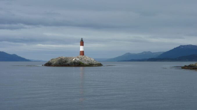Phare symbolisant la recherche de la vérité, situé sur un îlot rocheux, avec un paysage marin et montagneux en arrière-plan sous un ciel partiellement nuageux.