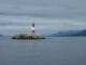 Phare symbolisant la recherche de la vérité, situé sur un îlot rocheux, avec un paysage marin et montagneux en arrière-plan sous un ciel partiellement nuageux.