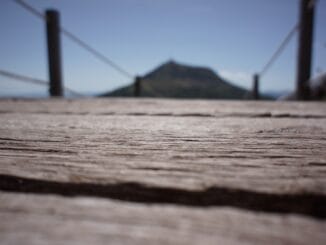 Un pont en bois en premier plan avec une mise au point rapprochée sur sa texture, menant au flou d'une montagne éloignée sous un ciel bleu, métaphore d'un voyage vers la sagesse.