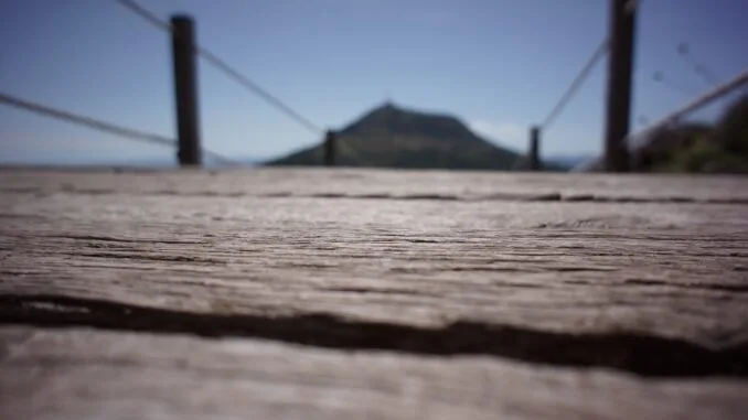 La Voie de la Sagesse : Perspective et Clarté Un pont en bois en premier plan avec une mise au point rapprochée sur sa texture, menant au flou d'une montagne éloignée sous un ciel bleu, métaphore d'un voyage vers la sagesse.